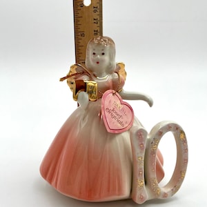 May include: A porcelain figurine of a girl in a pink dress holding a gift box that says "MY HERO". The figurine has a "10" next to it, with gold accents. A tag reads "Dakin Josef Originals".