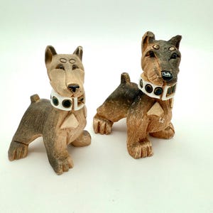 May include: Two wooden dog figurines. The dogs are brown and tan with white collars and black accents. The figurines are carved with a stylized design and have a whimsical appearance. The dogs are posed in a standing position.