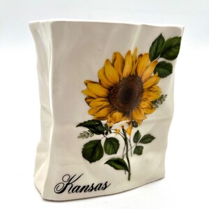 May include: Cream-colored ceramic vase with a sunflower design. The vase features a large yellow sunflower with a brown center, green leaves, and the word "Kansas" in script.