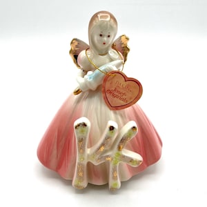 May include: A vintage porcelain figurine of a woman with angel wings, wearing a pink and white dress. The figurine holds a light blue object and has a heart-shaped tag that reads "DAKIN Josef Originals". The base features the letter "K".