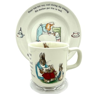 Puede incluir: Un juego de taza y plato de cerámica con ilustraciones de conejos en una escena de cuento. El plato tiene texto alrededor del borde. La taza tiene un asa y un posavasos transparente.