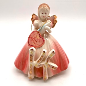 May include: A vintage porcelain figurine of a woman in a pink and white dress with gold accents. The figurine has a heart-shaped tag that reads "DAKIN Josef Originals". The figurine is holding a blue object and the number "14" in front.