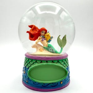May include: Ariel and Flounder snow globe. The figurine features Ariel with red hair and a green tail, embracing Flounder. The base is decorated with blue, green, and purple accents. The globe is clear.