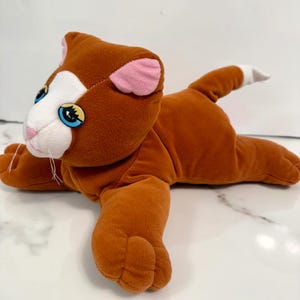 May include: A plush toy cat in a lying position. The cat is primarily brown with white and pink accents on the face and ears. It has blue eyes and a white-tipped tail. The toy is soft and cuddly.