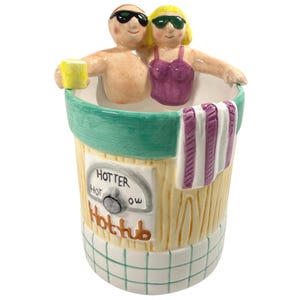 May include: A ceramic hot tub mug featuring two figures wearing sunglasses inside the tub. One figure holds a yellow drink. The exterior has a green rim, a purple and white striped towel, and a dial labeled HOTTER, HOT, and OW with the text Hottub.
