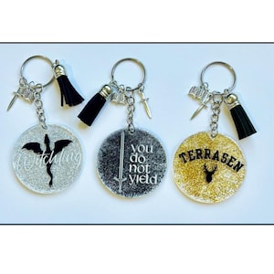 Puede incluir: Tres llaveros con colgantes redondos y brillantes. Cada llavero tiene un anillo plateado, una borla negra y abalorios. Los colgantes presentan texto y símbolos: un dragón, las palabras "you do not yield" y un ciervo.