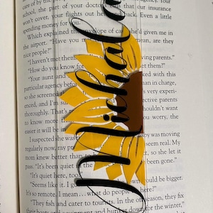 May include: Clear acrylic bookmark with a sunflower design. The bookmark features a yellow sunflower with black accents and the word "Smile" in a cursive font. A black tassel hangs from the top.