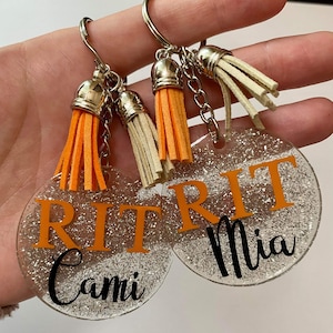 May include: Two clear, glittery acrylic keychains with silver keyrings and orange and cream tassels. Each keychain has the letters "RIT" in orange, with a name in black script below. One says "Cami" and the other says "Mia".