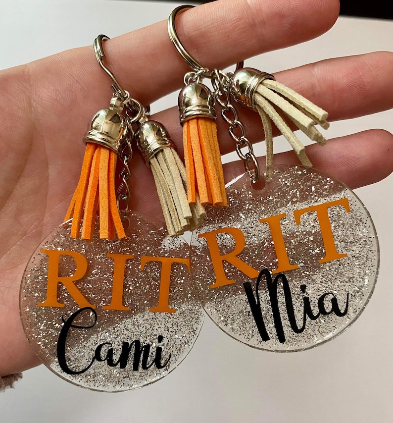May include: Two clear, glittery acrylic keychains with silver keyrings and orange and cream tassels. Each keychain has the letters "RIT" in orange, with a name in black script below. One says "Cami" and the other says "Mia".