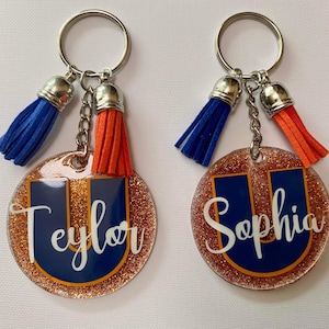 May include: Two personalized keychains with a glittery, round design. Each keychain features a navy blue letter "U" with a name in white script. One has a blue tassel, the other an orange tassel. Both have silver key rings.