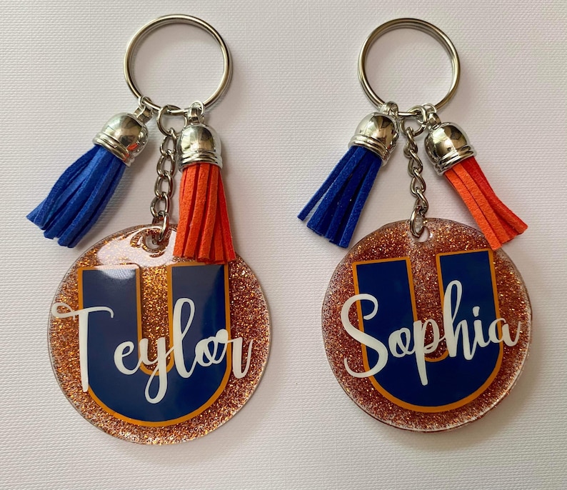 May include: Two personalized keychains with a glittery, round design. Each keychain features a navy blue letter "U" with a name in white script. One has a blue tassel, the other an orange tassel. Both have silver key rings.