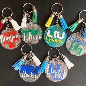 May include: A collection of personalized keychains with glittery, round acrylic pendants. Each keychain features a name or initials in various colors, paired with a matching tassel. The keychains are attached to silver key rings.
