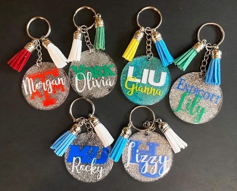 May include: A collection of personalized keychains with glittery, round acrylic pendants. Each keychain features a name or initials in various colors, paired with a matching tassel. The keychains are attached to silver key rings.