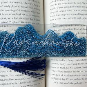 Puede incluir: Un marcapáginas de acrílico transparente con un diseño de purpurina azul y una borla azul. El marcapáginas presenta el nombre "Parzuchowski" en escritura blanca, con un diseño de olas.