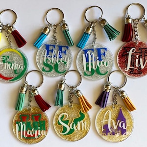 May include: A collection of personalized keychains with glittered circular pendants. Each keychain features a name or initials in various colors, with matching tassels. The keychains are silver-toned and have a variety of tassel colors.