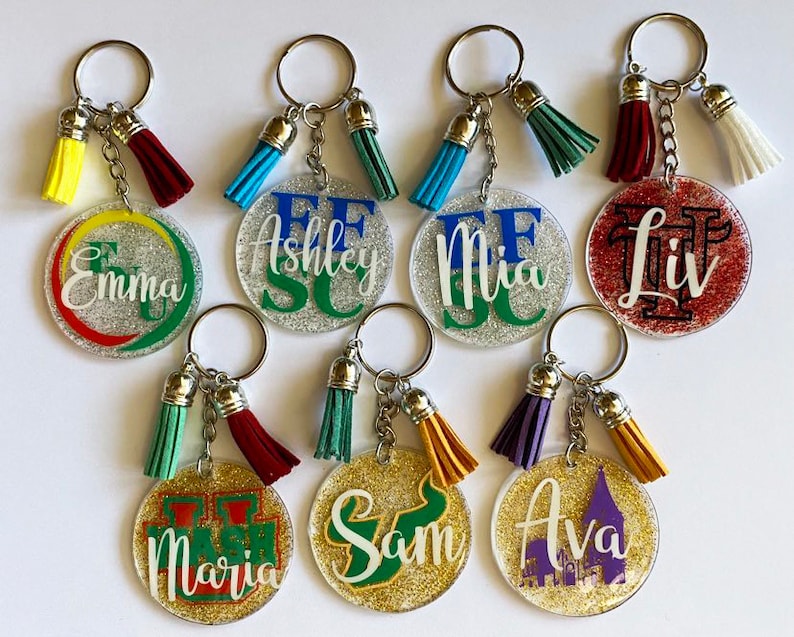 May include: A collection of personalized keychains with glittered circular pendants. Each keychain features a name or initials in various colors, with matching tassels. The keychains are silver-toned and have a variety of tassel colors.