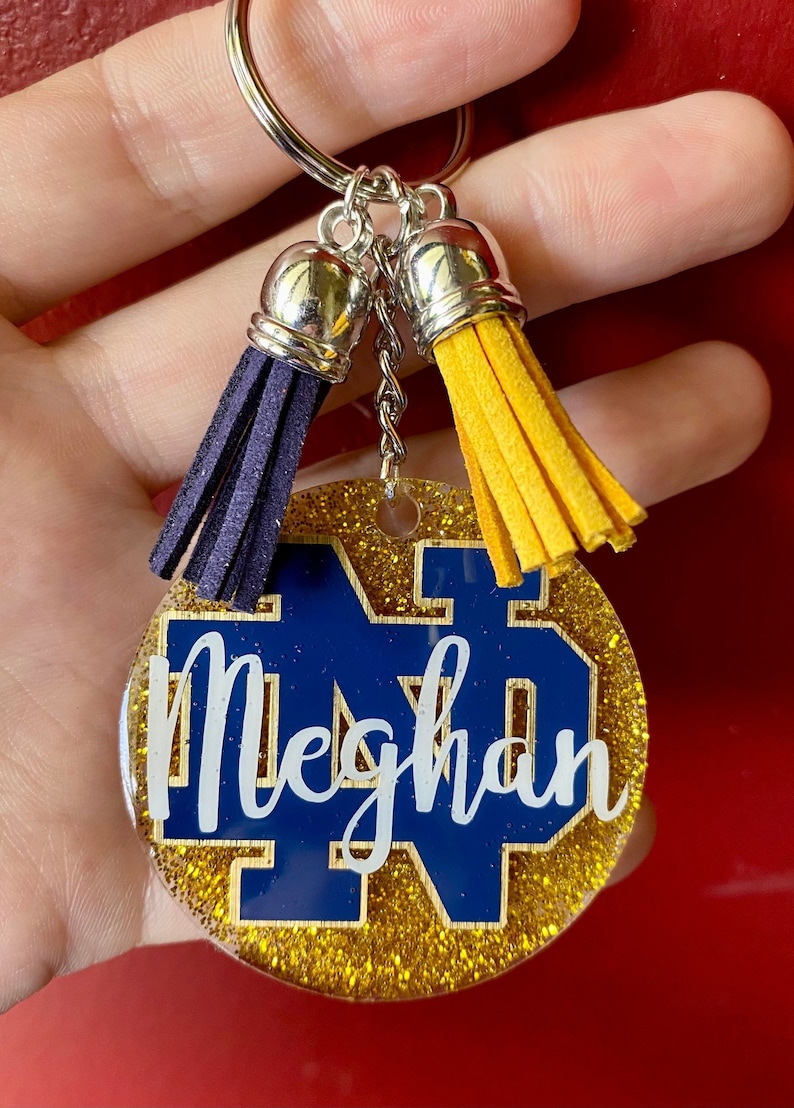 May include: A round keychain with a gold glitter border and a blue and gold design. The keychain has a blue and gold logo with the name "Meghan" in white script. It includes a silver keyring and two tassels, one blue and one yellow.