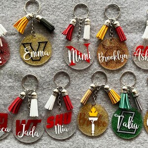 May include: A collection of personalized keychains with glittered backgrounds and various designs. Each keychain features a name or initial, a tassel, and a silver keyring. The keychains are in various colors, including red, gold, and green.