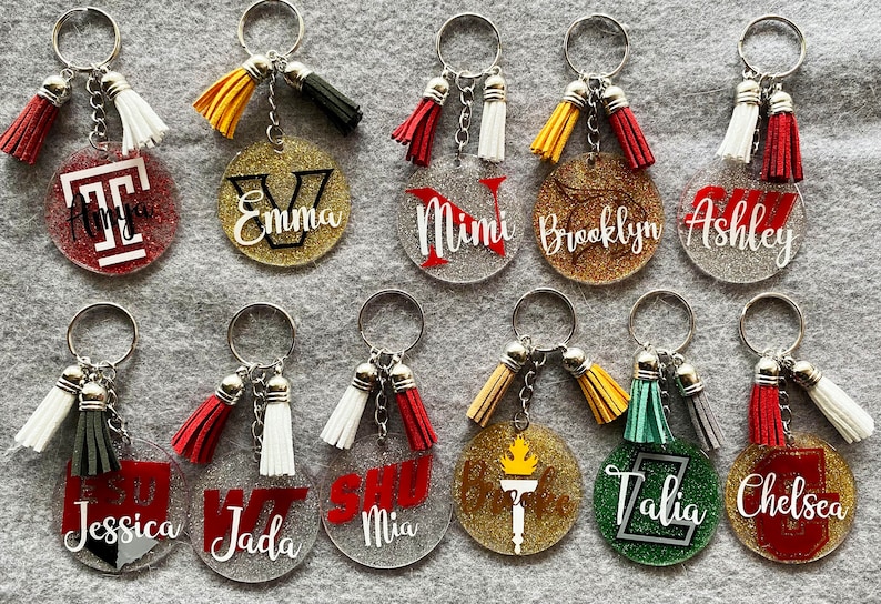 May include: A collection of personalized keychains with glittered backgrounds and various designs. Each keychain features a name or initial, a tassel, and a silver keyring. The keychains are in various colors, including red, gold, and green.