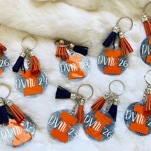 May include: Keychains with a silver glitter background, featuring an orange letter "I" and the text "DVM" with a year. Each keychain has a silver ring and two tassels, one navy blue and one orange.