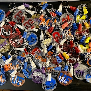 May include: A collection of personalized glitter keychains with names like "Haley", "Roni", and "Ally". Each keychain features a round or shaped charm with a name in a contrasting color, paired with a tassel in various colors. The keychains are silver-toned.