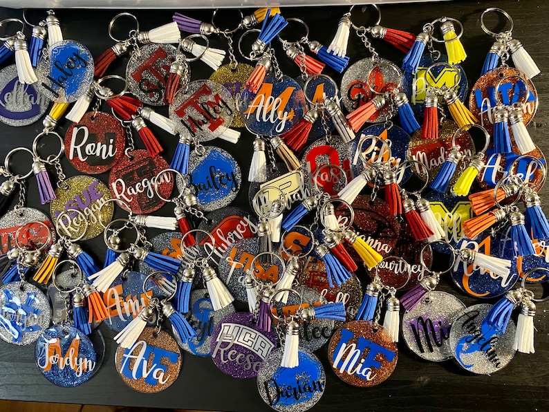 May include: A collection of personalized glitter keychains with names like "Haley", "Roni", and "Ally". Each keychain features a round or shaped charm with a name in a contrasting color, paired with a tassel in various colors. The keychains are silver-toned.