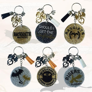 Może przedstawiać: Breloki z okrągłymi, błyszczącymi zawieszkami z napisami i zawieszkami w kształcie smoka. Breloki mają czarne, białe i beżowe frędzle. Teksty obejmują "BASGIATH WAR COLLEGE" i "SHOULD I GET THE WINGLEADER".