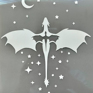May include: A white vinyl decal featuring a central dragon silhouette with expansive wings, surrounded by various small stars, circular dots, and a crescent moon. The design is presented on a smooth, light gray backing sheet, creating a high-contrast graphic.
