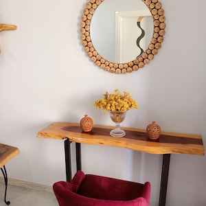 May include: A round mirror with a frame made of small, light-colored wooden pieces hangs above a wooden console table. The table has a live edge and black metal legs. A burgundy velvet chair sits in front of the table, and a vase of yellow flowers is on the table.