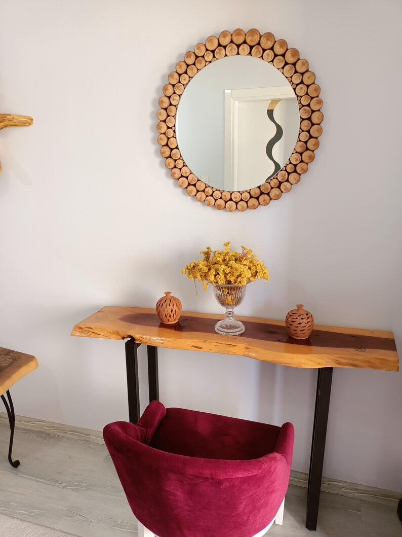 May include: A round mirror with a frame made of small, light-colored wooden pieces hangs above a wooden console table. The table has a live edge and black metal legs. A burgundy velvet chair sits in front of the table, and a vase of yellow flowers is on the table.