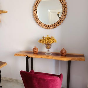 May include: A round mirror with a frame made of small, light-colored wooden discs. Below the mirror is a wooden console table with a live edge, supported by black metal legs. A burgundy velvet chair sits in front of the table.
