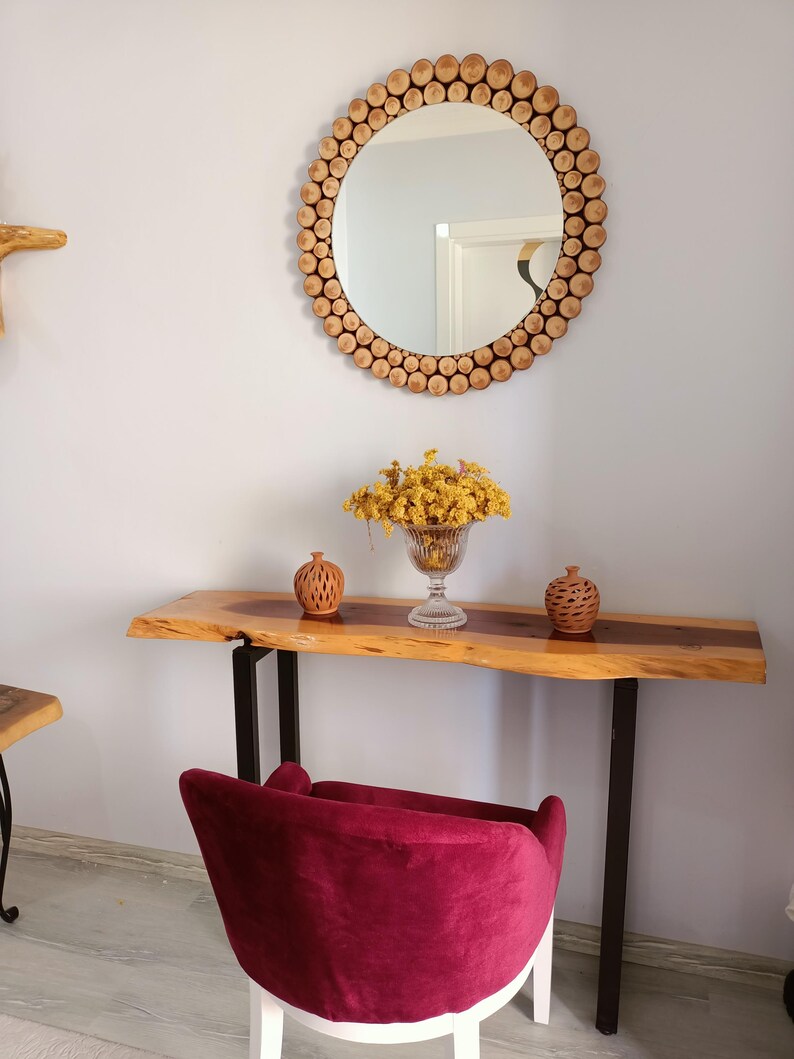 May include: A round mirror with a frame made of small, light-colored wooden discs. Below the mirror is a wooden console table with a live edge, supported by black metal legs. A burgundy velvet chair sits in front of the table.
