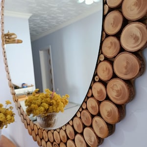 May include: Oval mirror with a decorative frame made of natural wood slices. The frame is composed of numerous circular wood pieces, creating a rustic and textured border around the reflective surface. The mirror is mounted on a white wall.