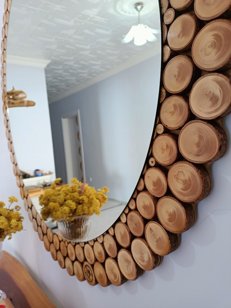 May include: Oval mirror with a decorative frame made of natural wood slices. The frame is composed of numerous circular wood pieces, creating a rustic and textured border around the reflective surface. The mirror is mounted on a white wall.