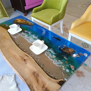 Op de afbeelding: Een houten salontafel met een blauw, groen en wit epoxyhars blad met een strandscène. De tafel heeft een natuurlijke houten rand en is omgeven door een wit tapijt.