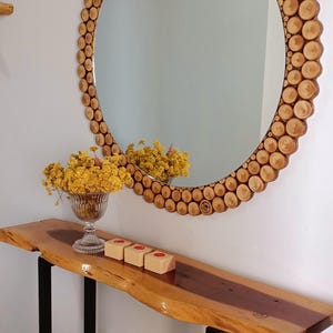 May include: A large round mirror with a wooden frame made of small, circular wood pieces. Below the mirror is a wooden console table with black metal legs. A glass vase with yellow flowers and three small wooden blocks with red circles are on the table.