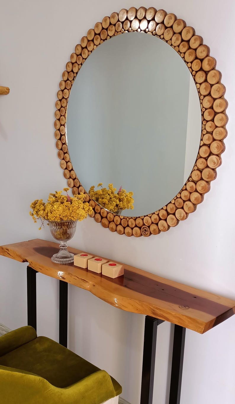 May include: A large round mirror with a wooden frame made of small, circular wood pieces. Below the mirror is a wooden console table with black metal legs. A glass vase with yellow flowers and three small wooden blocks with red circles are on the table.