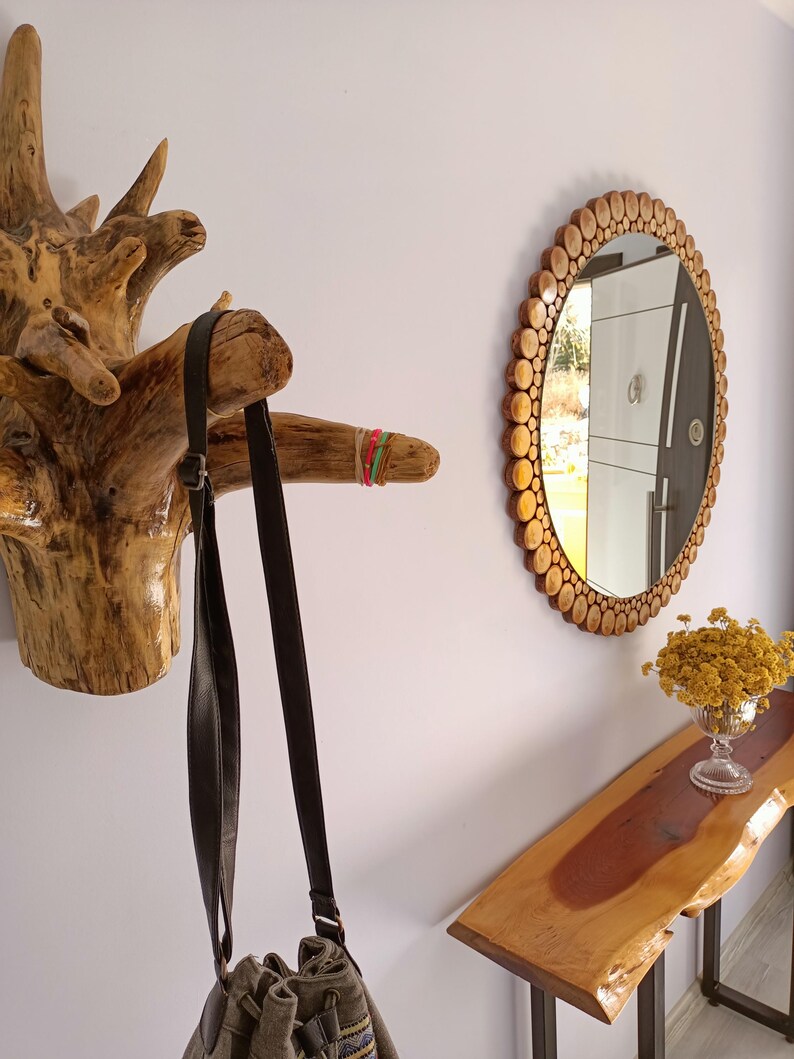 May include: A rustic entryway scene featuring a wooden coat rack, a round mirror with a wooden frame, and a live-edge console table. A dark brown shoulder bag hangs from the coat rack. A vase of yellow flowers sits on the table.
