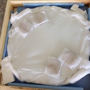 May include: A large, round, white object, possibly a mirror or artwork, is carefully packaged within a wooden crate. The object is wrapped in white protective material, with several smaller, round items secured on top. Blue foam padding lines the crate.