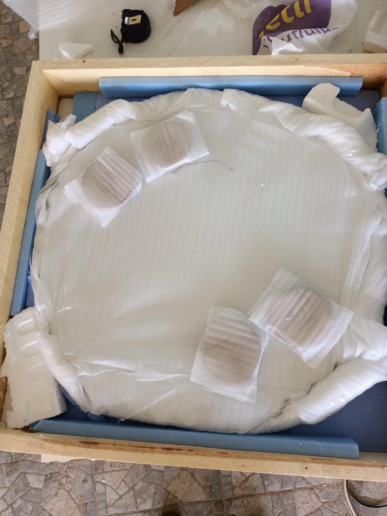 May include: A large, round, white object, possibly a mirror or artwork, is carefully packaged within a wooden crate. The object is wrapped in white protective material, with several smaller, round items secured on top. Blue foam padding lines the crate.