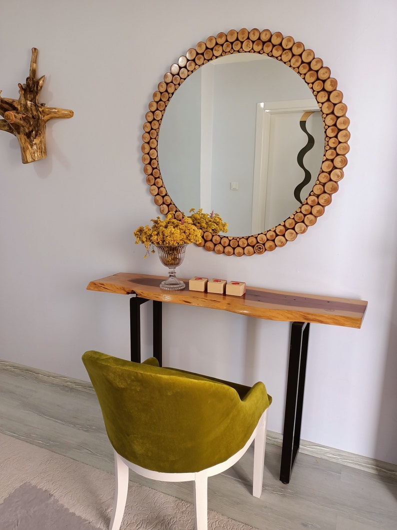 May include: A round mirror with a wooden frame made of small, circular wood pieces. Below the mirror is a wooden console table with black metal legs. A green velvet chair sits in front of the table. A decorative wooden piece hangs on the wall.
