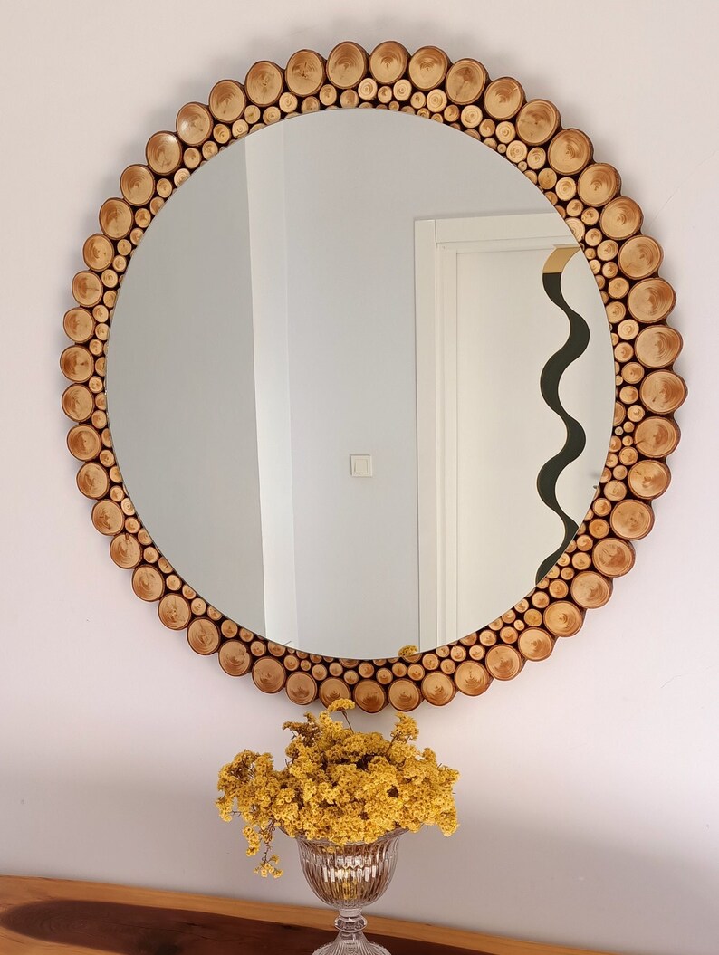May include: A round mirror with a decorative frame made of small, light brown wooden slices. The mirror is hanging on a white wall. A vase of yellow flowers sits below the mirror on a wooden surface.