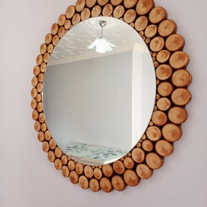 May include: Round mirror with a natural wood frame. The frame is made of small, circular wood pieces arranged around the mirror's edge. The mirror reflects a room with a light fixture and a patterned surface.