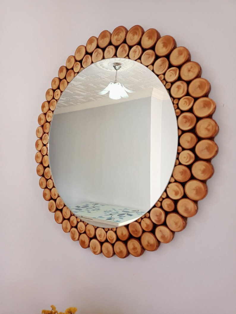 May include: Round mirror with a natural wood frame. The frame is made of small, circular wood pieces arranged around the mirror's edge. The mirror reflects a room with a light fixture and a patterned surface.