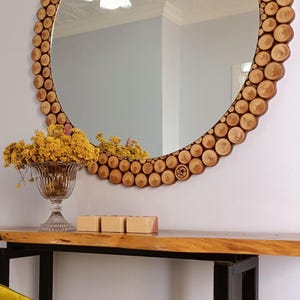 May include: A round mirror with a wooden frame made of small, circular wood pieces. The mirror is mounted on a wall above a wooden console table. A glass vase with yellow flowers and three small wooden boxes are on the table.