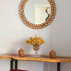 May include: A round mirror with a frame made of small, light brown wooden pieces. Below the mirror is a wooden console table with a glass vase of yellow flowers and two small terracotta vases. A red chair is in the foreground.