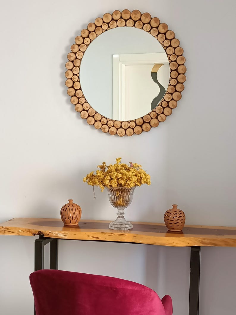 May include: A round mirror with a frame made of small, light brown wooden pieces. Below the mirror is a wooden console table with a glass vase of yellow flowers and two small terracotta vases. A red chair is in the foreground.