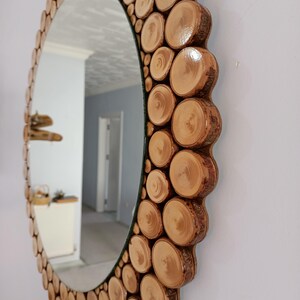 May include: An oval mirror with a decorative frame made of natural wood slices. The frame is composed of numerous circular wood pieces, creating a textured, rustic aesthetic. The mirror reflects a room with white walls and a doorway.