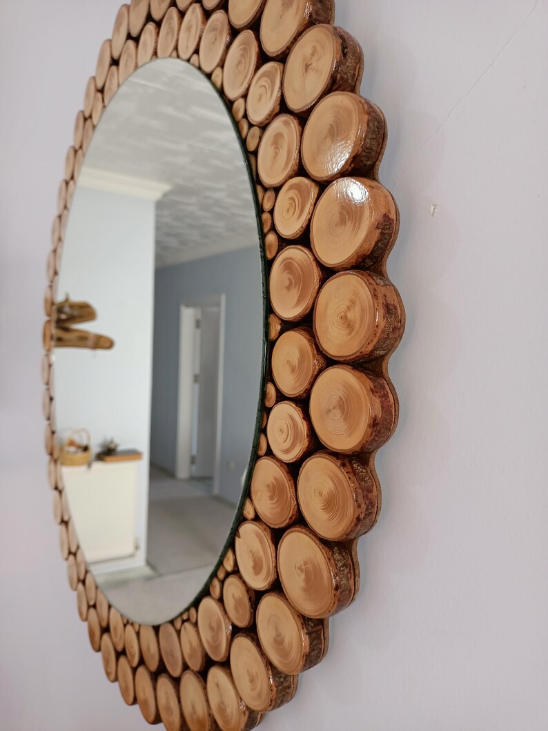 May include: An oval mirror with a decorative frame made of natural wood slices. The frame is composed of numerous circular wood pieces, creating a textured, rustic aesthetic. The mirror reflects a room with white walls and a doorway.