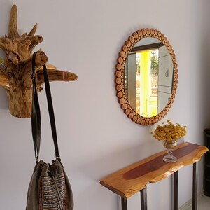 May include: A rustic entryway scene featuring a wooden coat rack, a round mirror with a wooden frame, and a live-edge console table. A green and patterned drawstring bag hangs from the coat rack. A vase of yellow flowers sits on the table.